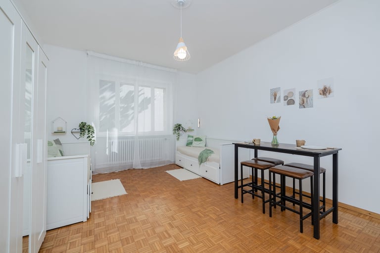 Bright minimalist room with wooden herringbone floor, black bar table with stools, window with radiator, and potted plants