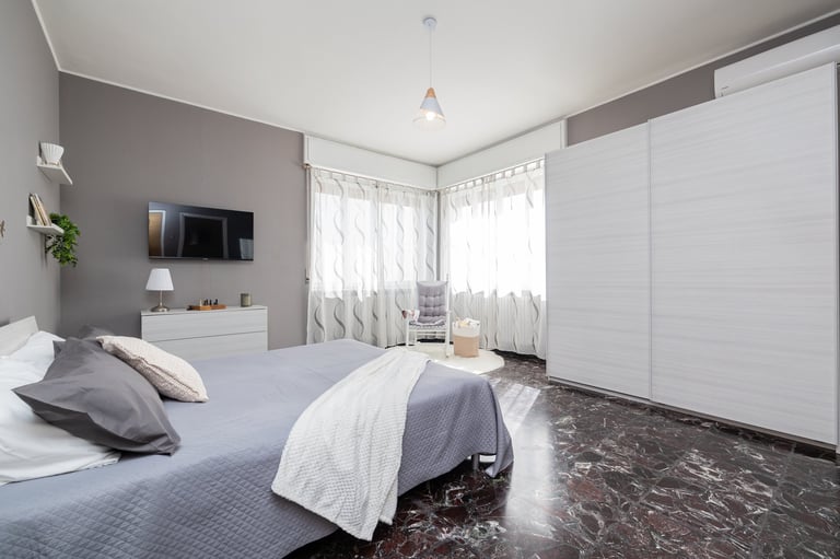 Modern bedroom with gray accent wall, white bedding, TV, and floor-to-ceiling white wardrobe with natural light from windows