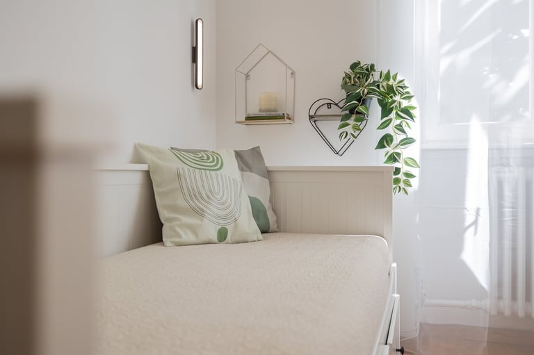 Bright bedroom corner with white bed, geometric patterned pillows, wall-mounted plant hanger, and decorative wire wall art