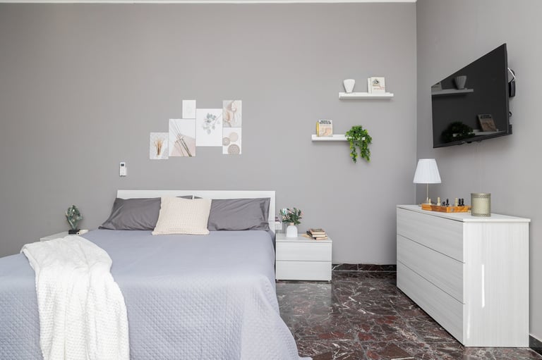 Modern bedroom with blue bed, gray pillows, white nightstand, white dresser, mounted TV, wall art, and potted plants